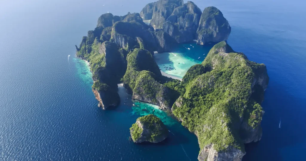 Majestic rock formations resembling small mountains emerging from the ocean near a tropical island in Thailand, surrounded by calm blue waters.